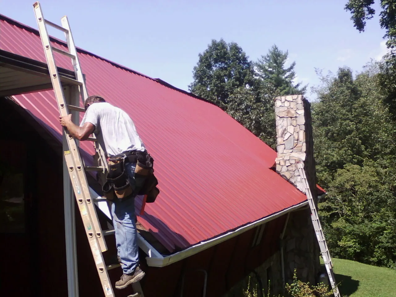 Roofer on a ladder beside a red metal roof and stone chimney — Soffit & Fascia Repair by Roofing Brothers in Wheatland